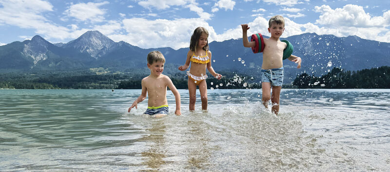 Drei kleine Kinder planschen im See, im Hintergrund die Bergkulisse.