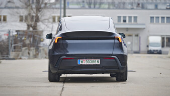Heck eines dunkelgrauen Tesla Model Y, das auf einer Straße vor einem hellgrauen Gebäude steht.