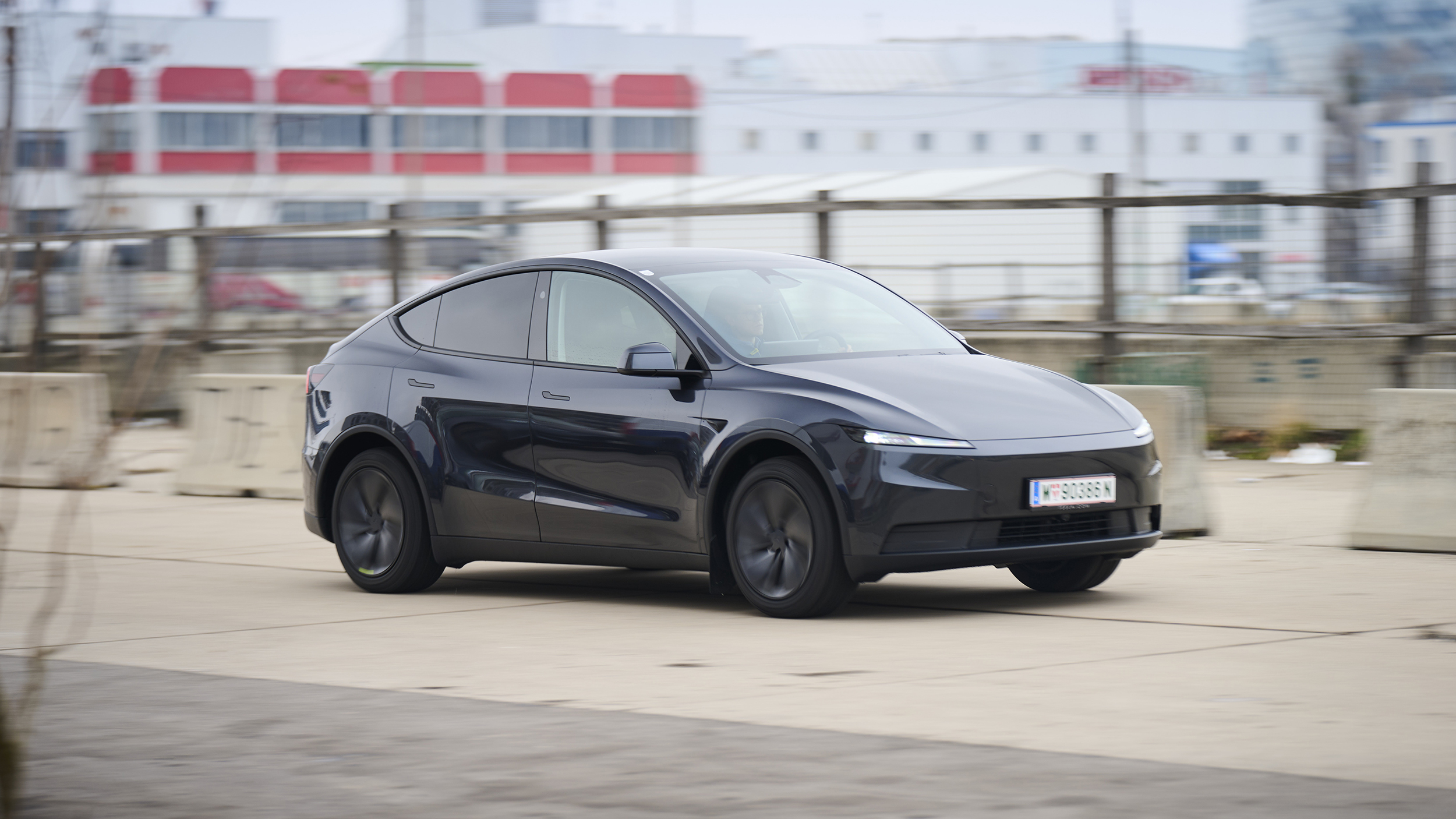 Ein dunkelgrauer Tesla Model Y fährt auf einer Straße vor einem grauen Gebäude.