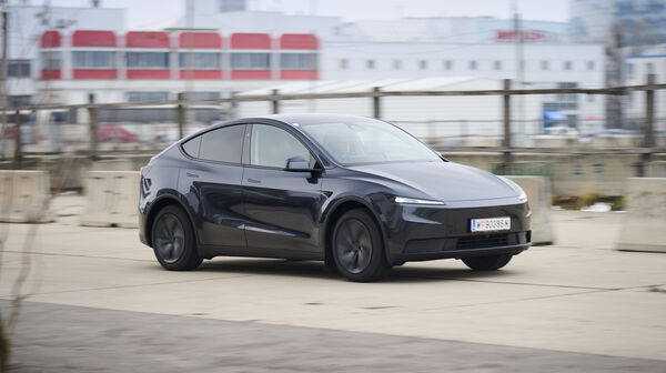 Ein dunkelgrauer Tesla Model Y fährt auf einer Straße vor einem grauen Gebäude.