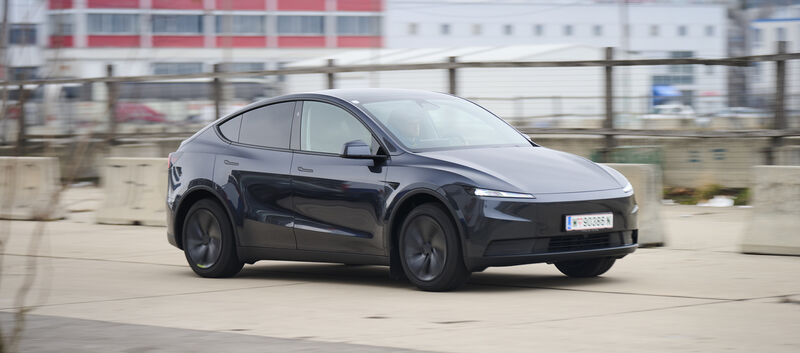 Ein dunkelgrauer Tesla Model Y fährt auf einer Straße vor einem grauen Gebäude.