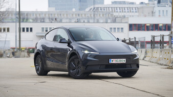 Front eines dunkelgrauen Tesla Model Y, das vor einem grauen Gebäude.