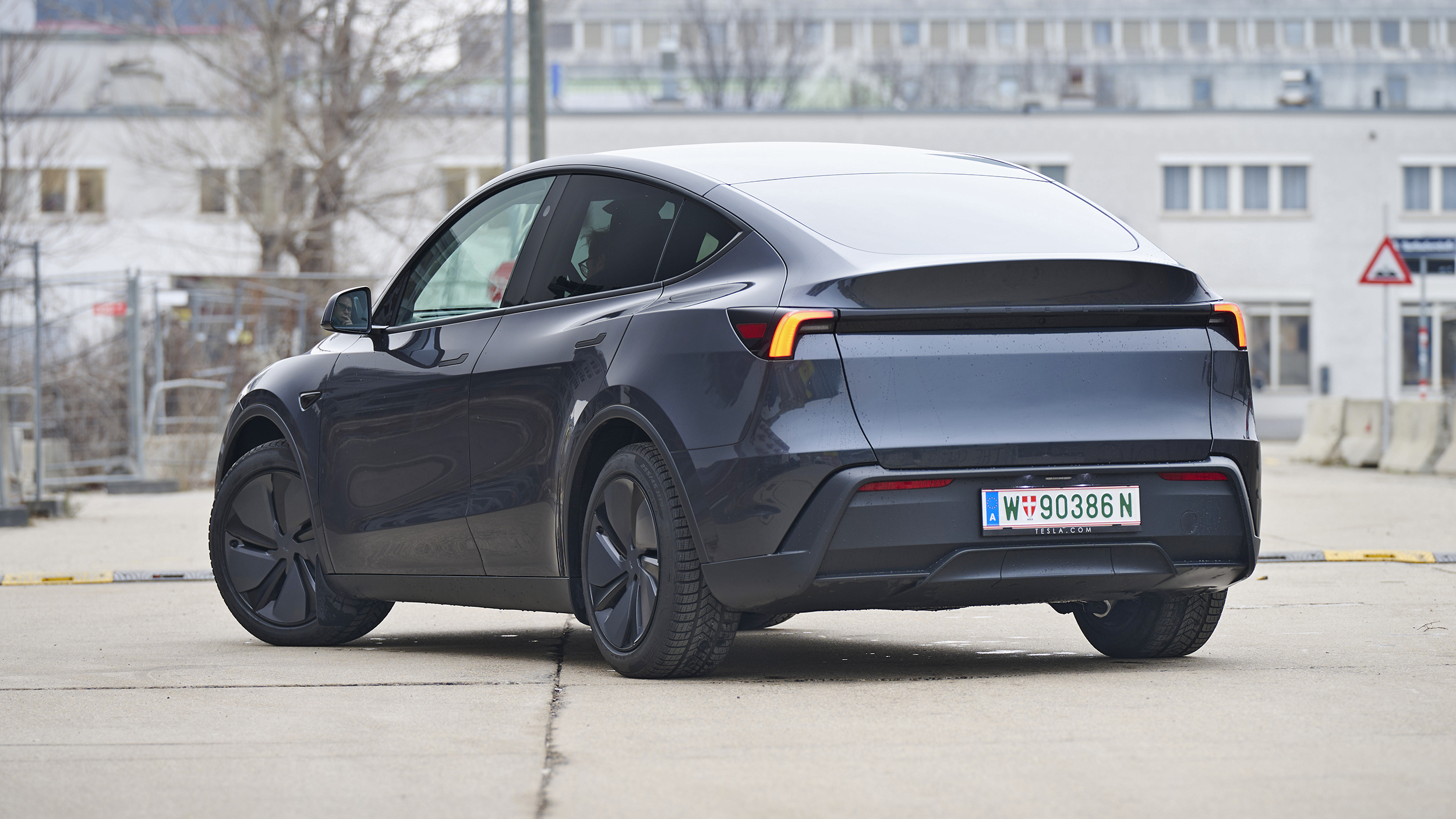 Heck eines dunkelgrauen Tesla Model Y, das auf einer Straße vor einem hellgrauen Gebäude steht.