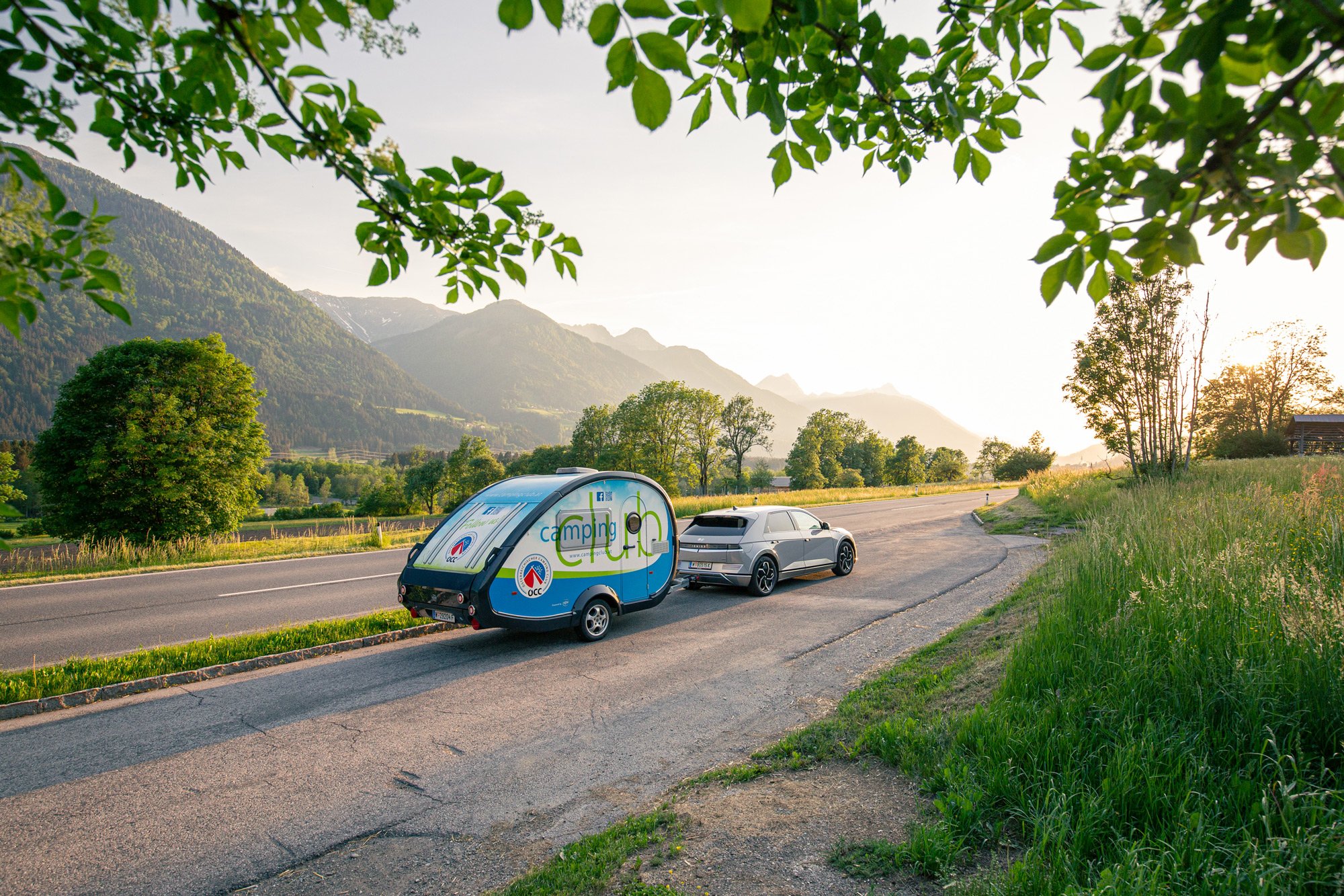 Es ist ein Straße bei Sonnenuntergang zu sehen. Es fährt ein Auto mit dem ÖCC-Campinganhänger.