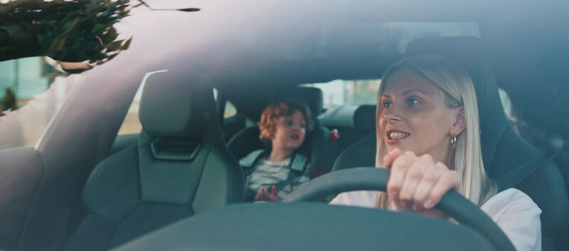 Eine blonde Frau am Steuer eines Autos, am Rücksitz sitzt ein Kind.