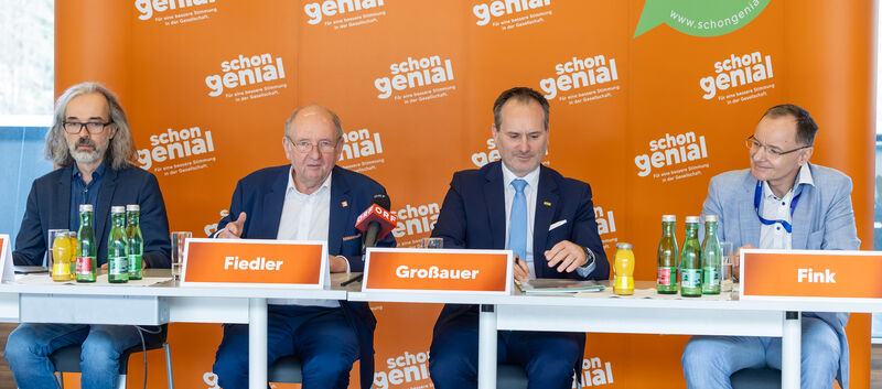 Vier Herren im Anzug sitzen an einem Tisch und geben eine Pressekonferenz