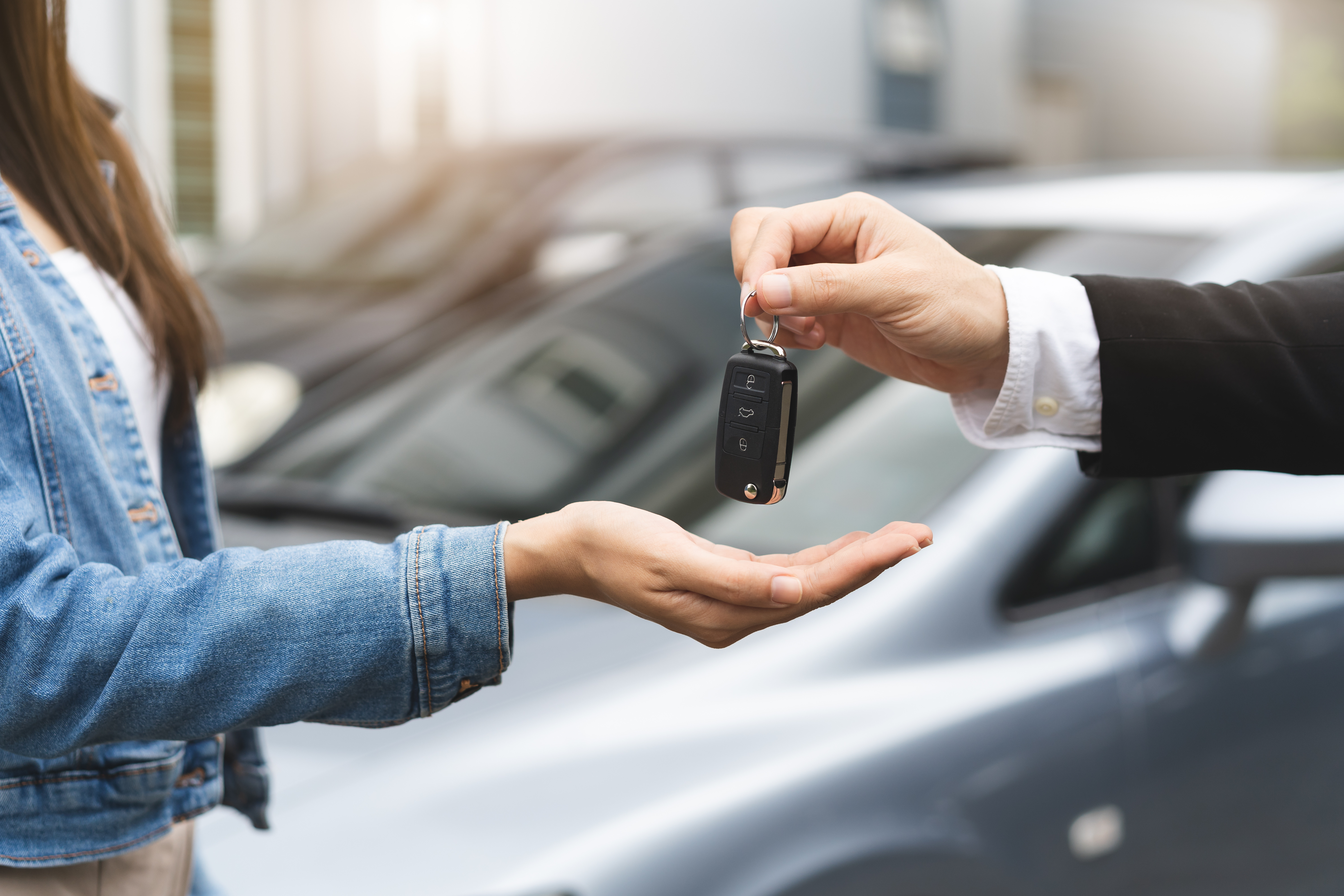 Eine Hand hält einen Autoschlüssel und gibt ihn einer anderen Person weiter. Im Hintergrund stehen zwei Autos.