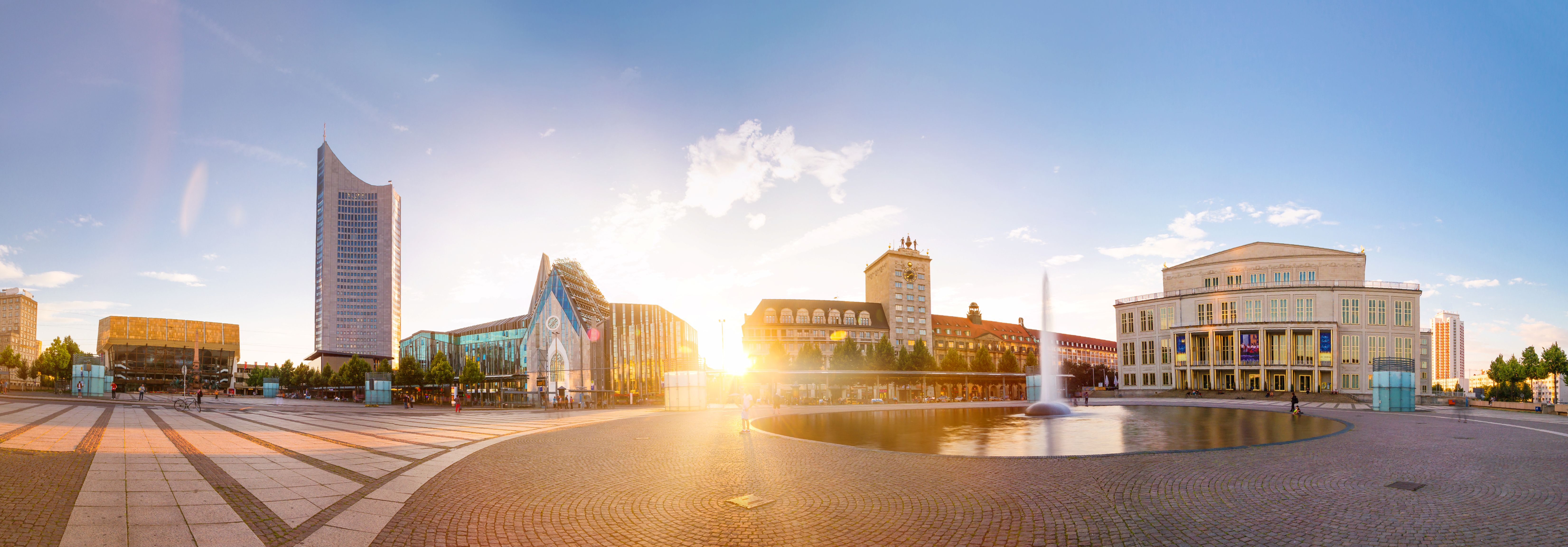 Panoramablick auf den Leipziger Augustusplatz bei Sonnenuntergang mit modernen und historischen Gebäuden, einem Brunnen und einem Kopfsteinpflasterplatz. Helle, ruhige Atmosphäre.