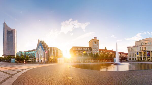 Panoramablick auf den Leipziger Augustusplatz bei Sonnenuntergang mit modernen und historischen Gebäuden, einem Brunnen und einem Kopfsteinpflasterplatz. Helle, ruhige Atmosphäre.