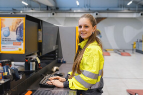 Es ist eine junge Frau zu sehen, die an einem Computer steht und arbeitet.