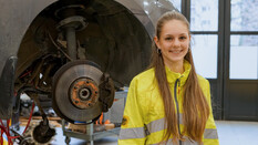 Es ist eine junge Frau zu sehen, die in einer Werkstatt steht.