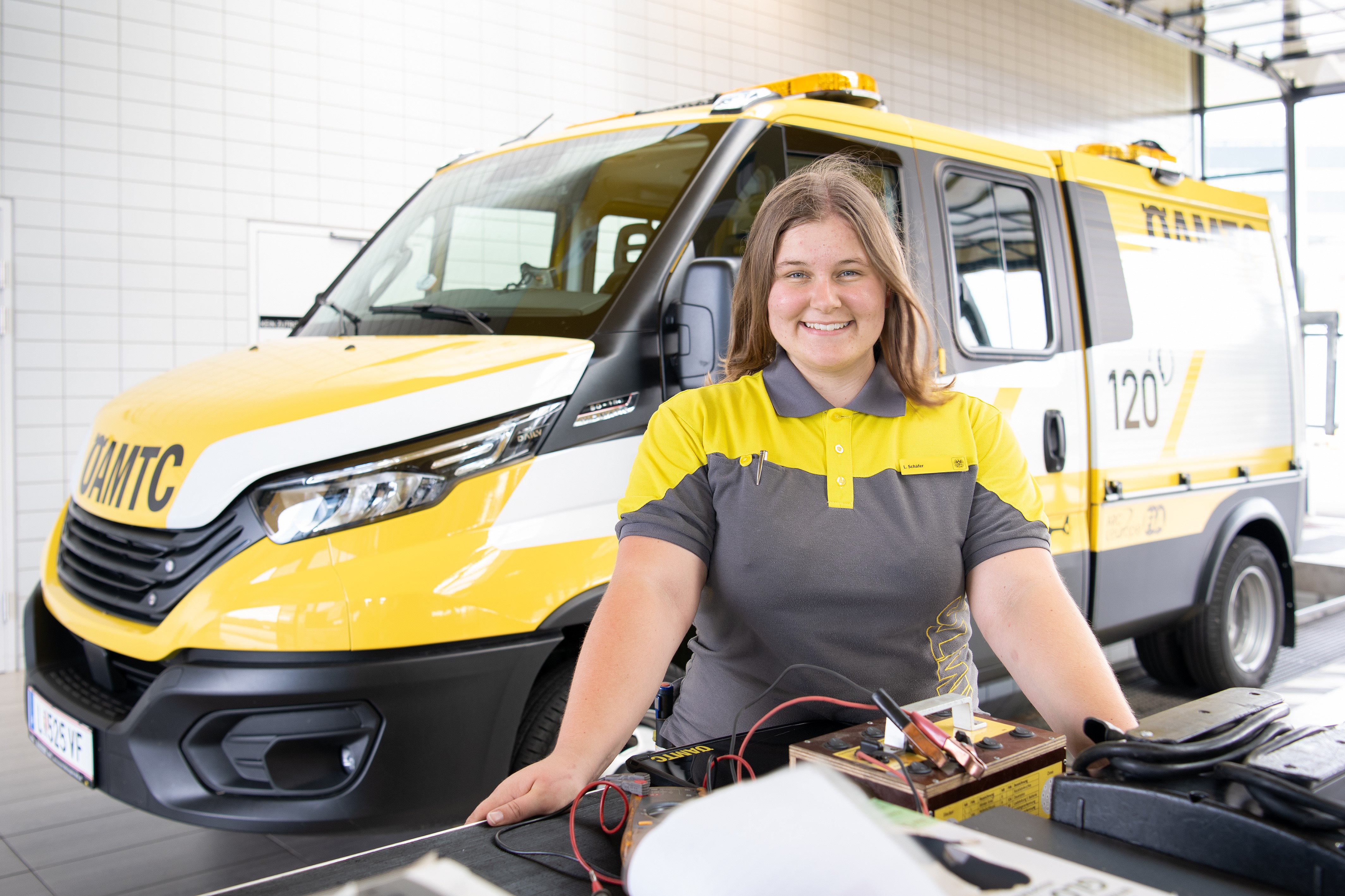 Es ist eine Frau in ÖAMTC-Kleidung zu sehen. Hinter ihr sieht man ein Pannenfahrzeug.