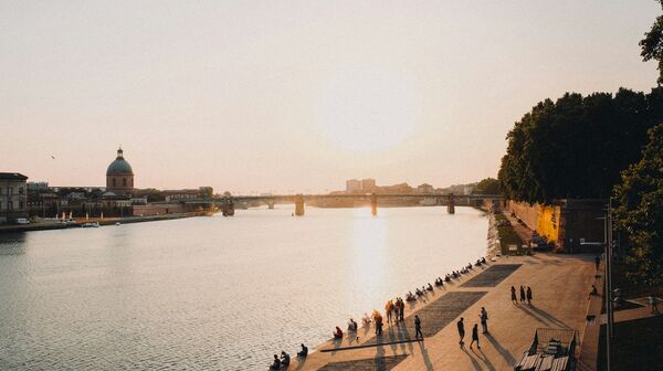 Sonnenuntergang an der Garonne in Toulouse, an deren Uferpromenade gemächlich Menschen spazieren und sitzen. Die Silhouette einer Kuppel und einer Brücke in der Ferne unterstreicht die friedliche Szenerie.