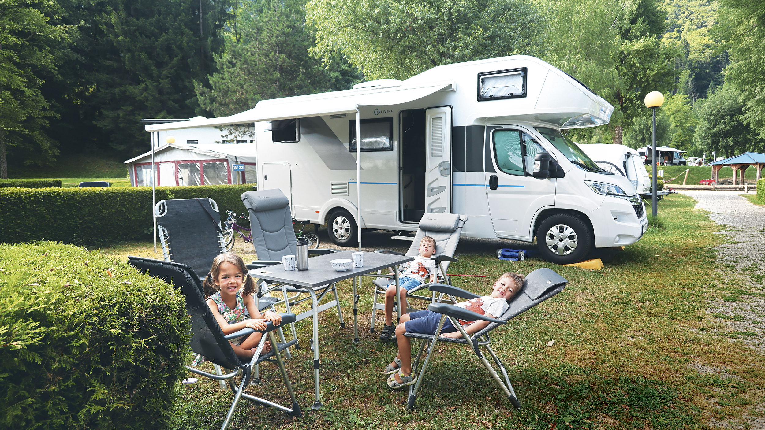 Drei kleine Kinder sitzen am Tisch vor ihrem Wohnmobil.