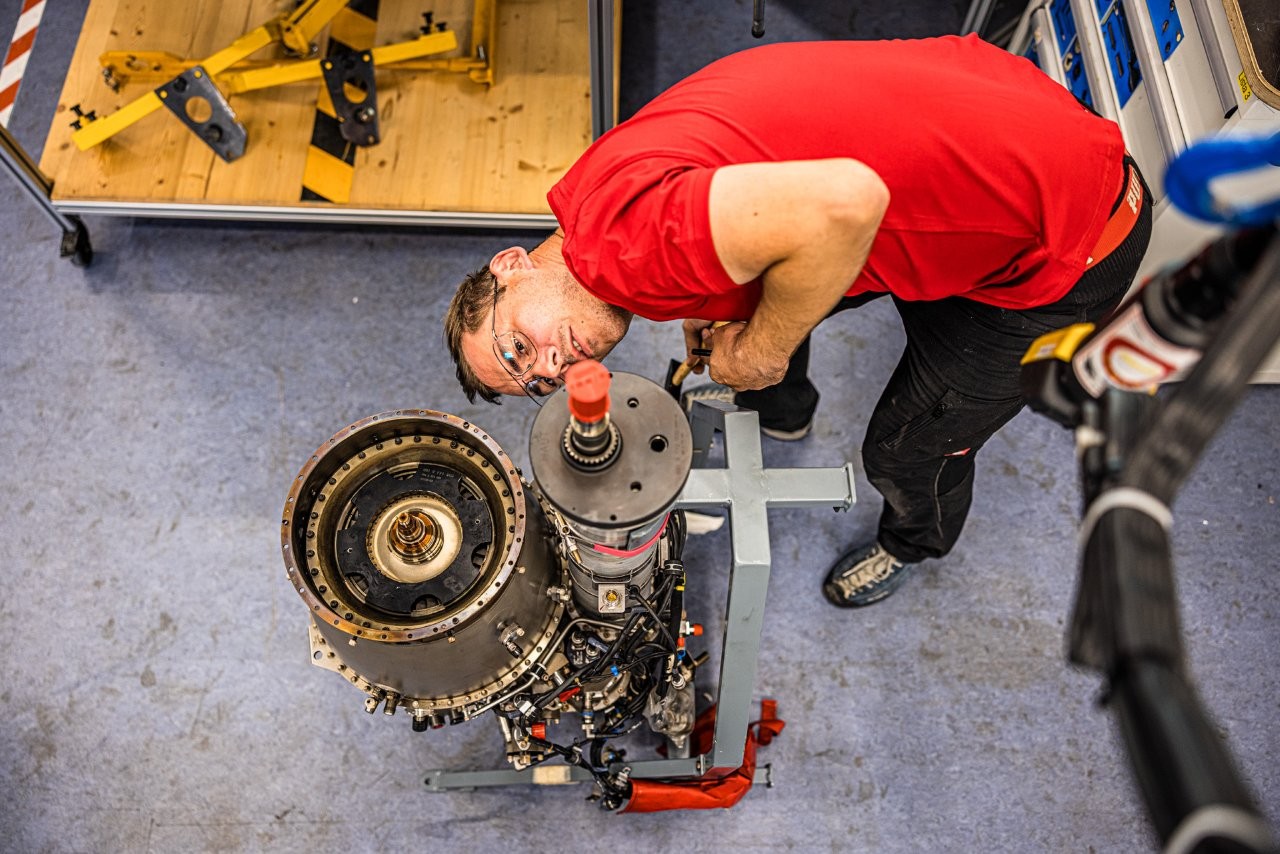 Auf dem Bild sieht man einen jungen Mann im roten T-Shirt, der eine Hubschrauber Turbine inspiziert.