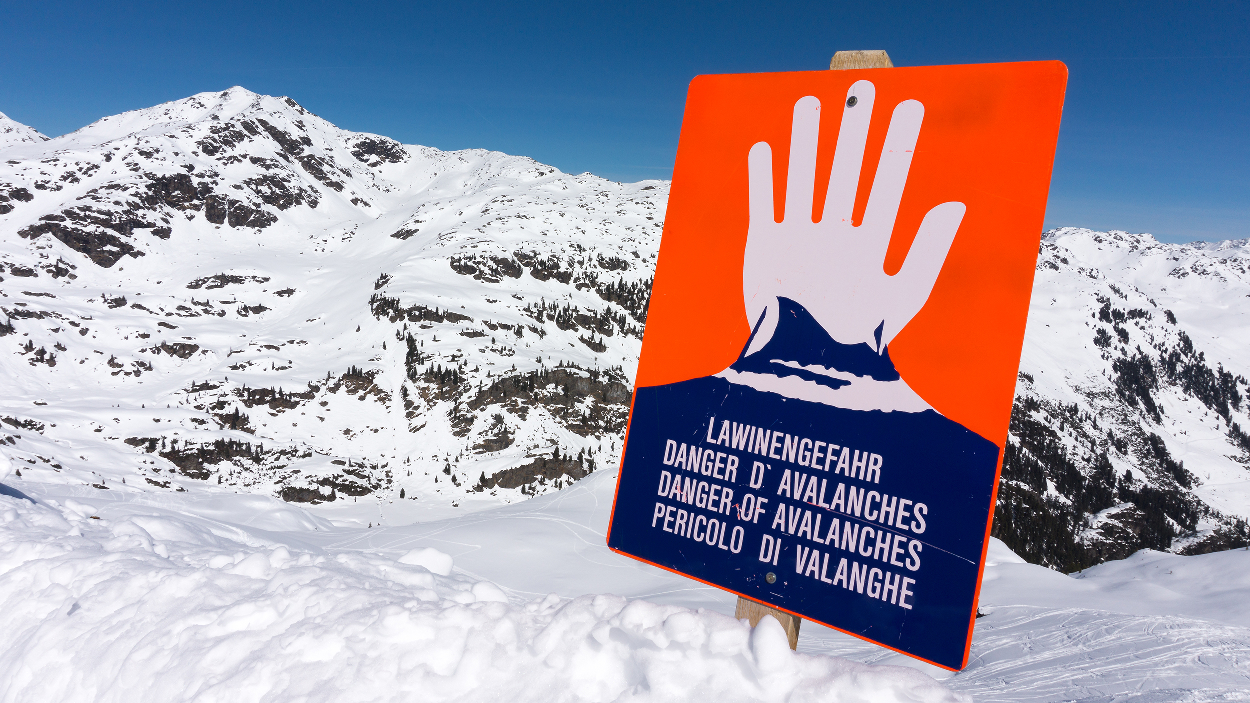 Ein rotes Lawinengefahr-Schild, man sieht eine weiße Hand, die "Halt" signalisiert. Dahinter sind die Berge zu sehen.