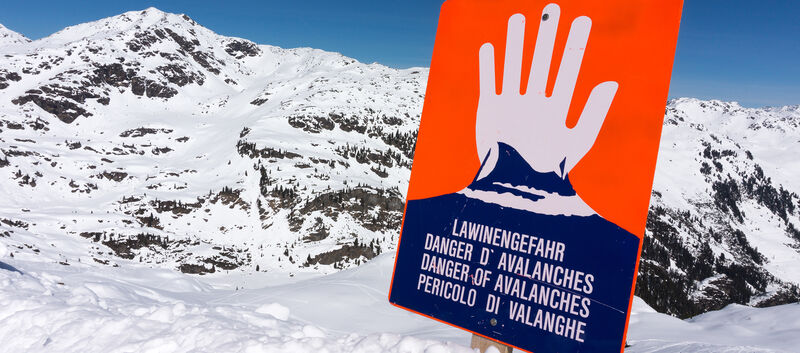 Ein rotes Lawinengefahr-Schild, man sieht eine weiße Hand, die "Halt" signalisiert. Dahinter sind die Berge zu sehen.