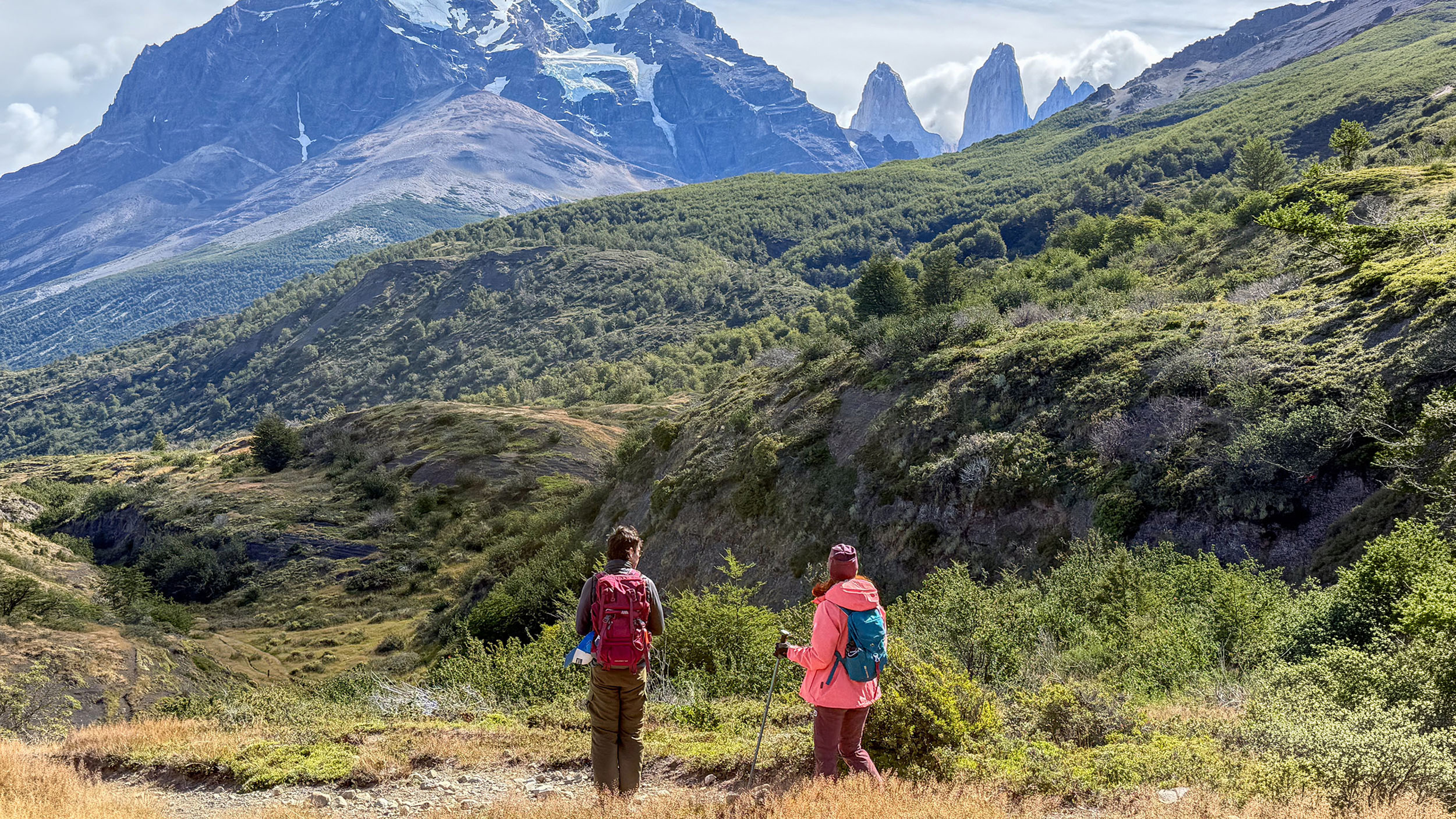 zwei Wanderer blicken von einer Lichtung auf die drei Granit-Berge Tres Torres del Paine, die hinter iem Hügel hervorschauen.