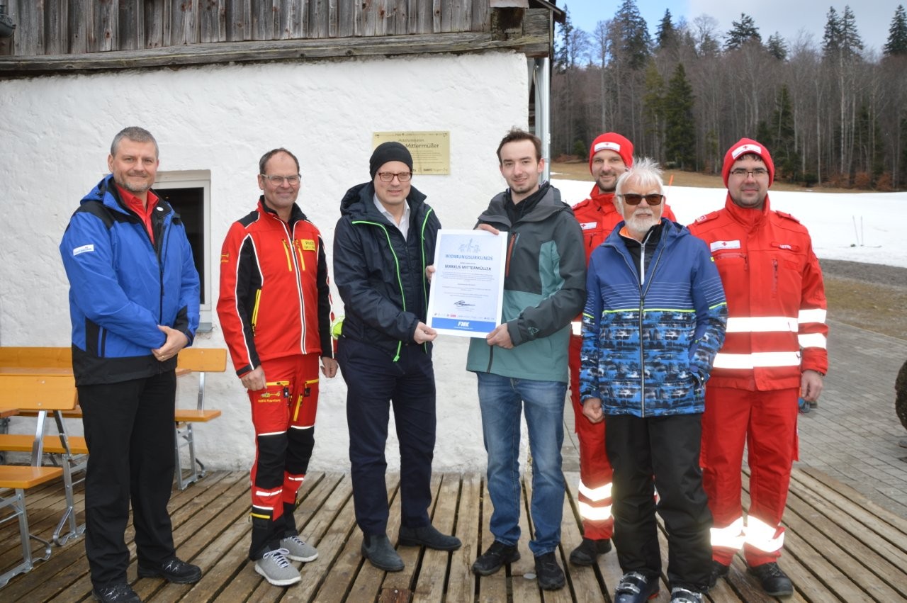 Auf dem Bild sieht man sieben Männer vor einem Haus. Der Mittlere hält eine Urkunde in der Hand. Drei von Ihnen haben rote Uniformen unterschiedlicher Rettungsdienste an. Die anderen sind winterlich gekleidet.