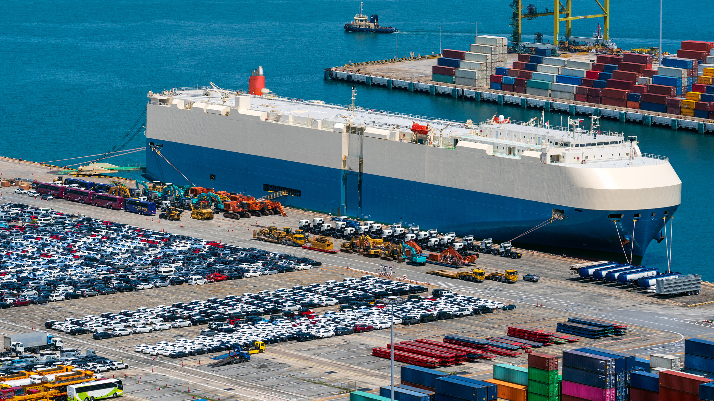 Tausende Autos auf einem Parkplatz an einem Hafen, ein Transportschiff liegt vor Anker.