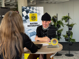 Das Bild zeigt Jan Wunderlich im interview. Er sitzt bei einem Tisch, neben ihm steht sein Preis. Ihm gegenüber sitzt eine Frau, die man nur von hinten sieht.