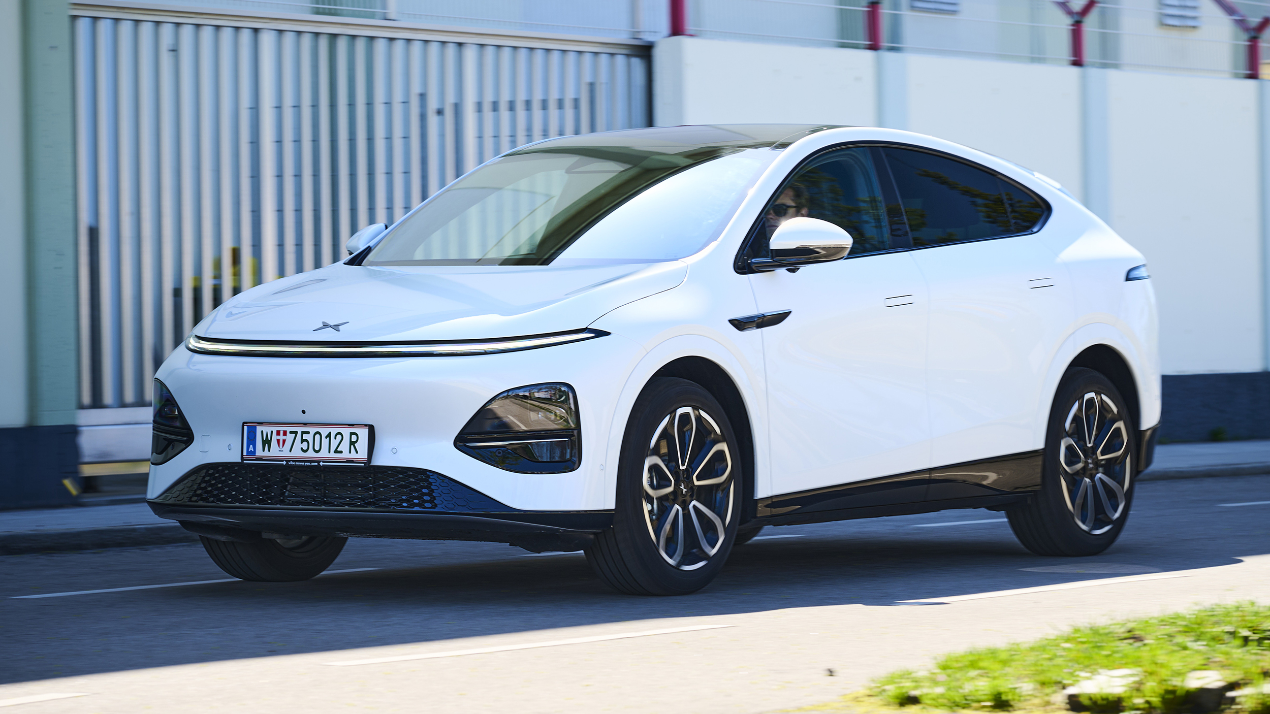 Ein weißer Xpeng G6 fährt auf einer Nebenstraße vor einem Gebäude.