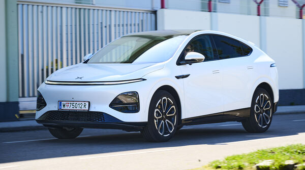 Ein weißer Xpeng G6 fährt auf einer Nebenstraße vor einem Gebäude.