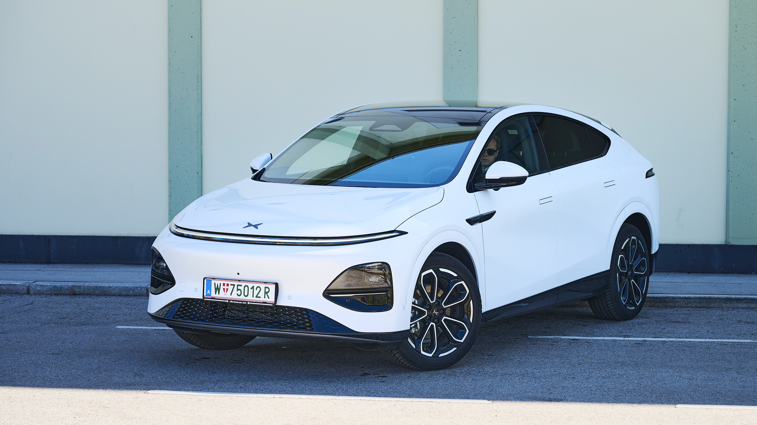Ein weißer Xpeng G6 steht quer auf der Straße vor einer leicht grünen Wand.