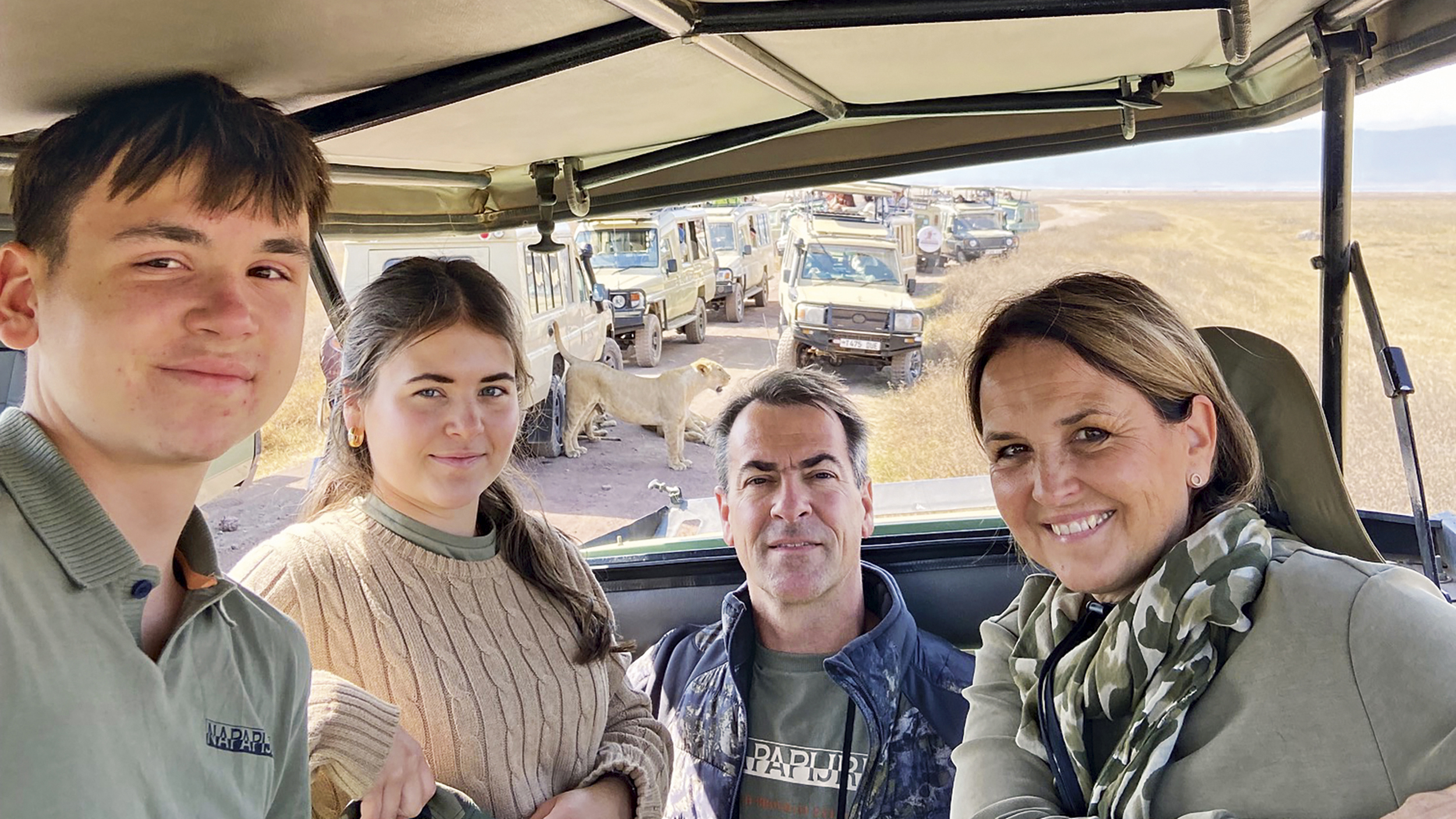 Familie mit zwei Teenager-Kindern steht im offenen Safari-Jeep, dahinter sieht man einen Löwen.