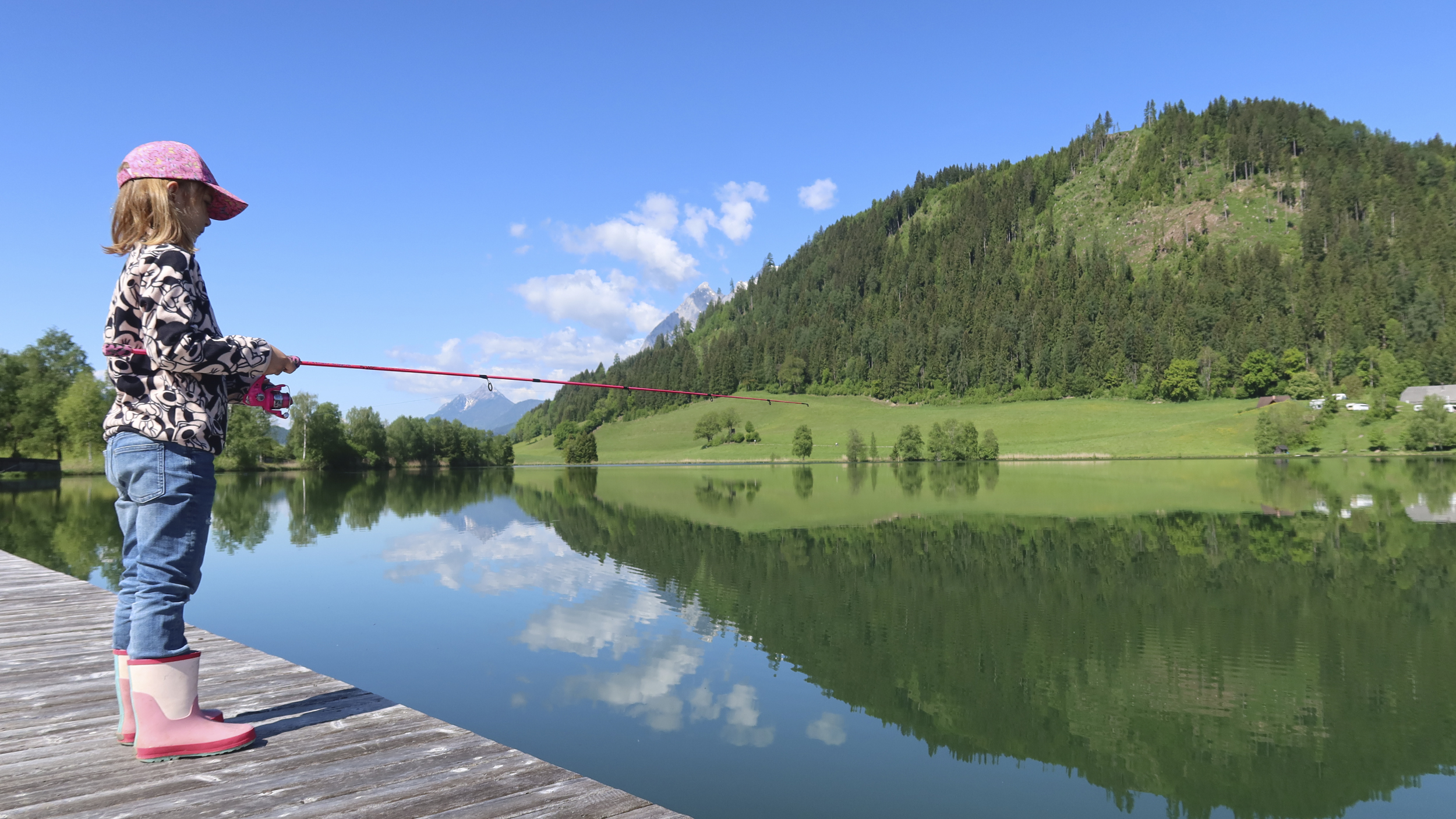 Ein Mädchen angelt von einem Steg weg, im Hintergrund bewaldete Berge.
