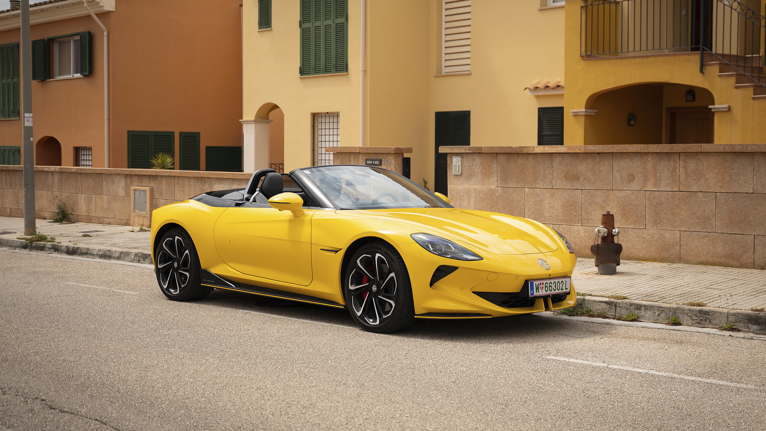 Ein gelber MG Cyberster steht auf einem Parkplatz vor einem orangenen Haus.