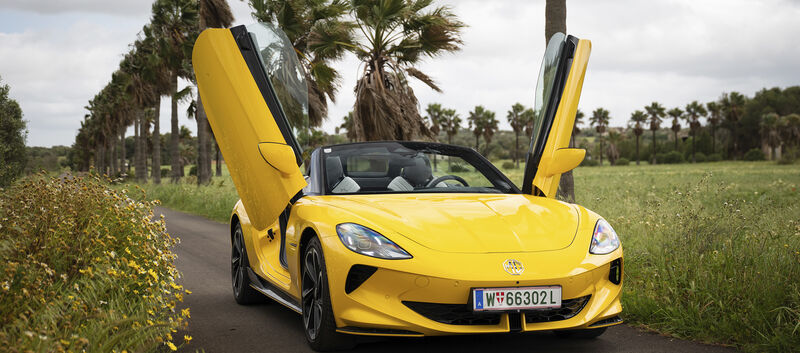 Ein gelber MG Cyberster steht mit geöffneten Scherentüren auf einer Straße, die von Wiese umgeben ist.