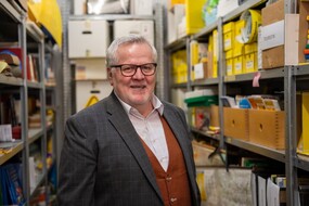 Auf dem Bild sieht man einen Mann mit Brille, Sakko und weißem Hemd zwischen Regalen voll mit Kartons stehen.