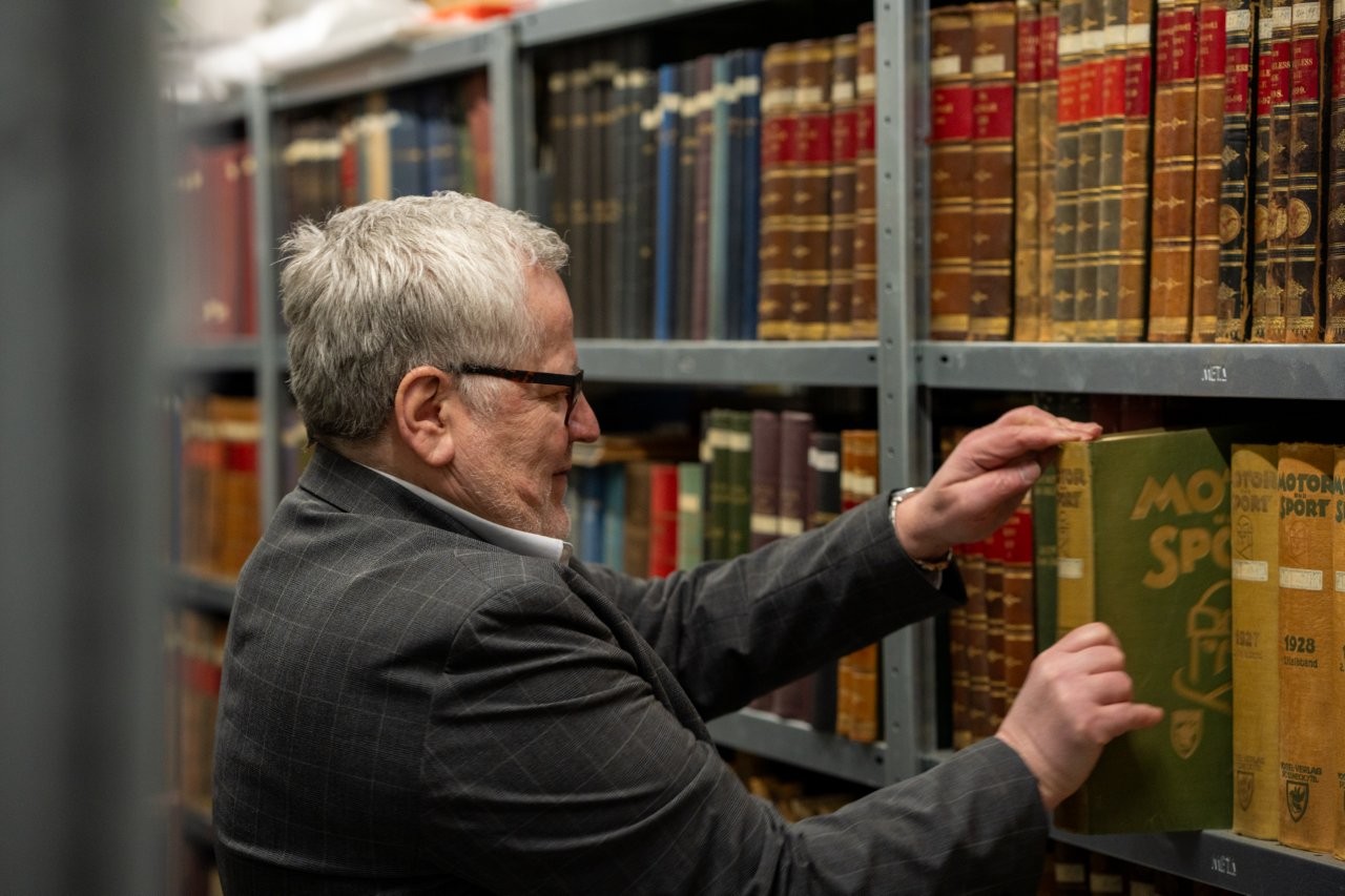 Auf dem Bild sieht man einen Mann mit Brille, der ein altes Buch aus einem Regal zieht.