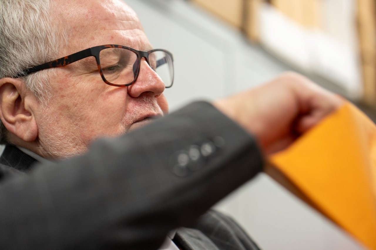 Auf dem Bild sieht man die Nahaufnahme eines Gesichts eines Mannes der konzentriert etwas aus einem gelben Kuvert nimmt.