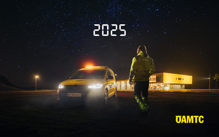 Ein gelbes Pannenhilfeauto steht vor einem großen Gebäude. Ein Pannenhelfer mit gelber Warnkleidung geht auf das Fahrzeug zu. Es ist Nacht und Sterne sind am Himmel sichtbar. 2025 steht als Schriftzug am Himmel.