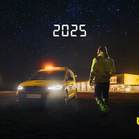 Ein gelbes Pannenhilfeauto steht vor einem großen Gebäude. Ein Pannenhelfer mit gelber Warnkleidung geht auf das Fahrzeug zu. Es ist Nacht und Sterne sind am Himmel sichtbar. 2025 steht als Schriftzug am Himmel.