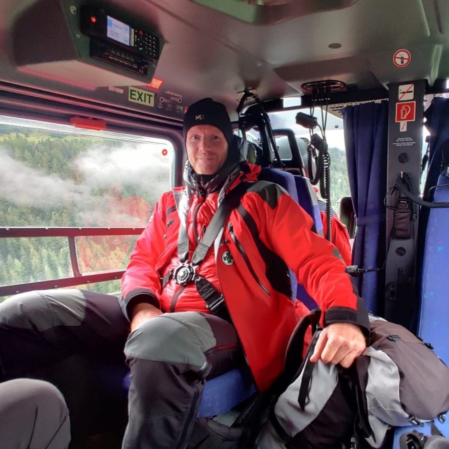 Auf dem Bild sieht man einen Bergretter in rotem Gewand, der in einem Hubschrauber sitzt.