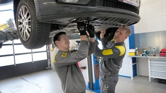 Auf dem Bild sieht man zwei Mitarbeiter des ÖAMTC, die unter einem Fahrzeug stehen, dass auf einer Hebebühne steht.