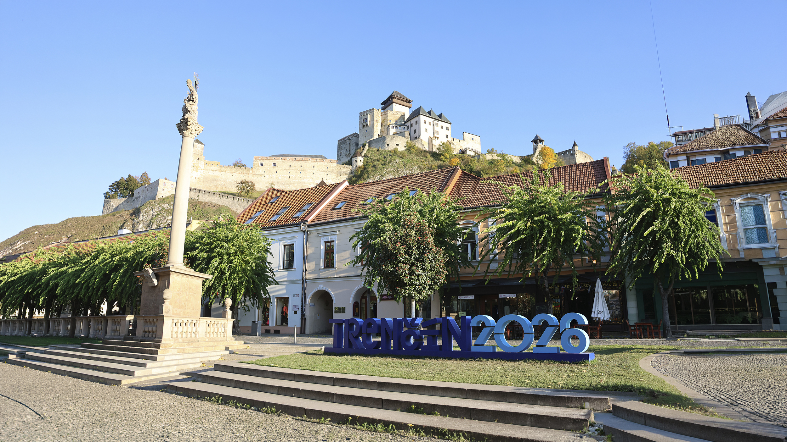 Der Hauptplatz mit einer Säule und einem blauen großen Schriftzug "Trencin 2026", dahinter auf einem Hügel eine Burg.