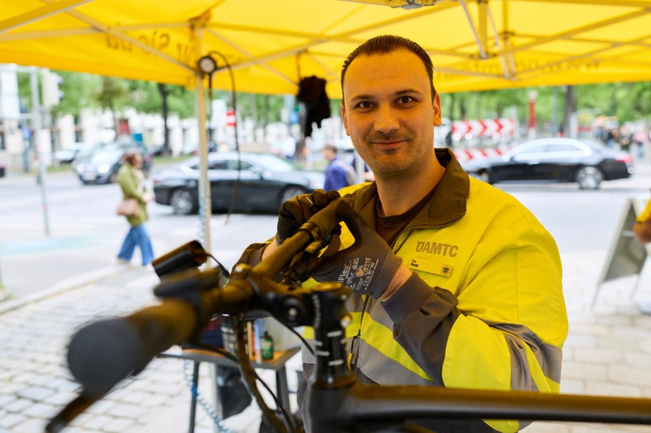 Auf dem Bild sieht man einen Mitarbeiter des ÖAMTC hinter einem Fahrrad. Der Mann schaut in die Kamera und hat eine gelb/graue Jacke an.