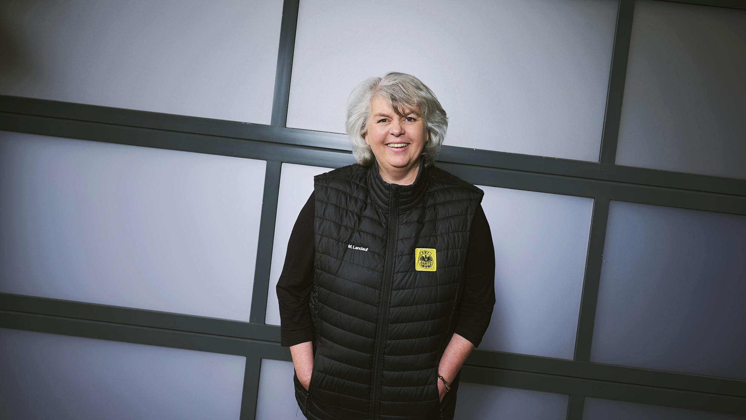 Eine Frau mit grauen Haaren und schwarz gekleidet steht vor einer Wand.