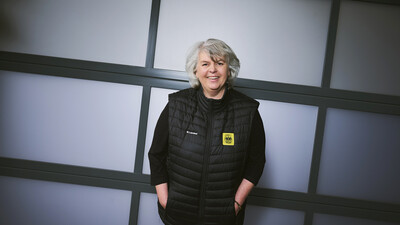 Eine Frau mit grauen Haaren und schwarz gekleidet steht vor einer Wand.