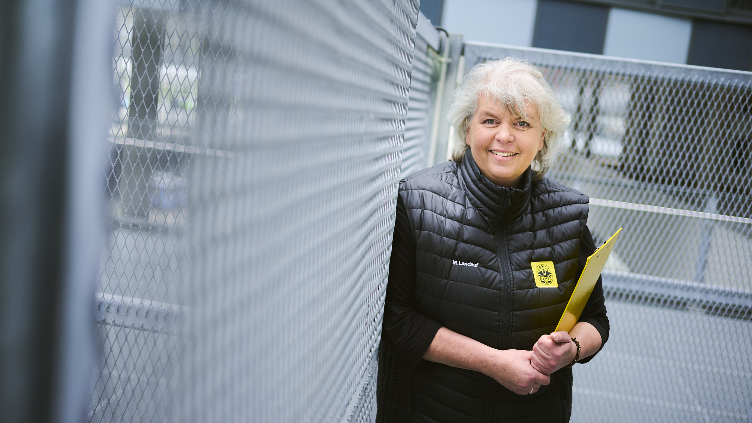 Eine Frau mit grauen Haaren und schwarz gekleidet steht vor einem Zaun. Sie hält ein Klemmbrett und lacht.