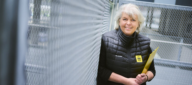 Eine Frau mit grauen Haaren und schwarz gekleidet steht vor einem Zaun. Sie hält ein Klemmbrett und lacht.