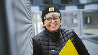 Eine Frau mit grauen Haaren, Haube und schwarz gekleidet steht vor einem Zaun und lacht. Michaela Landauf hält ein gelbes Klemmbrett in der Hand.