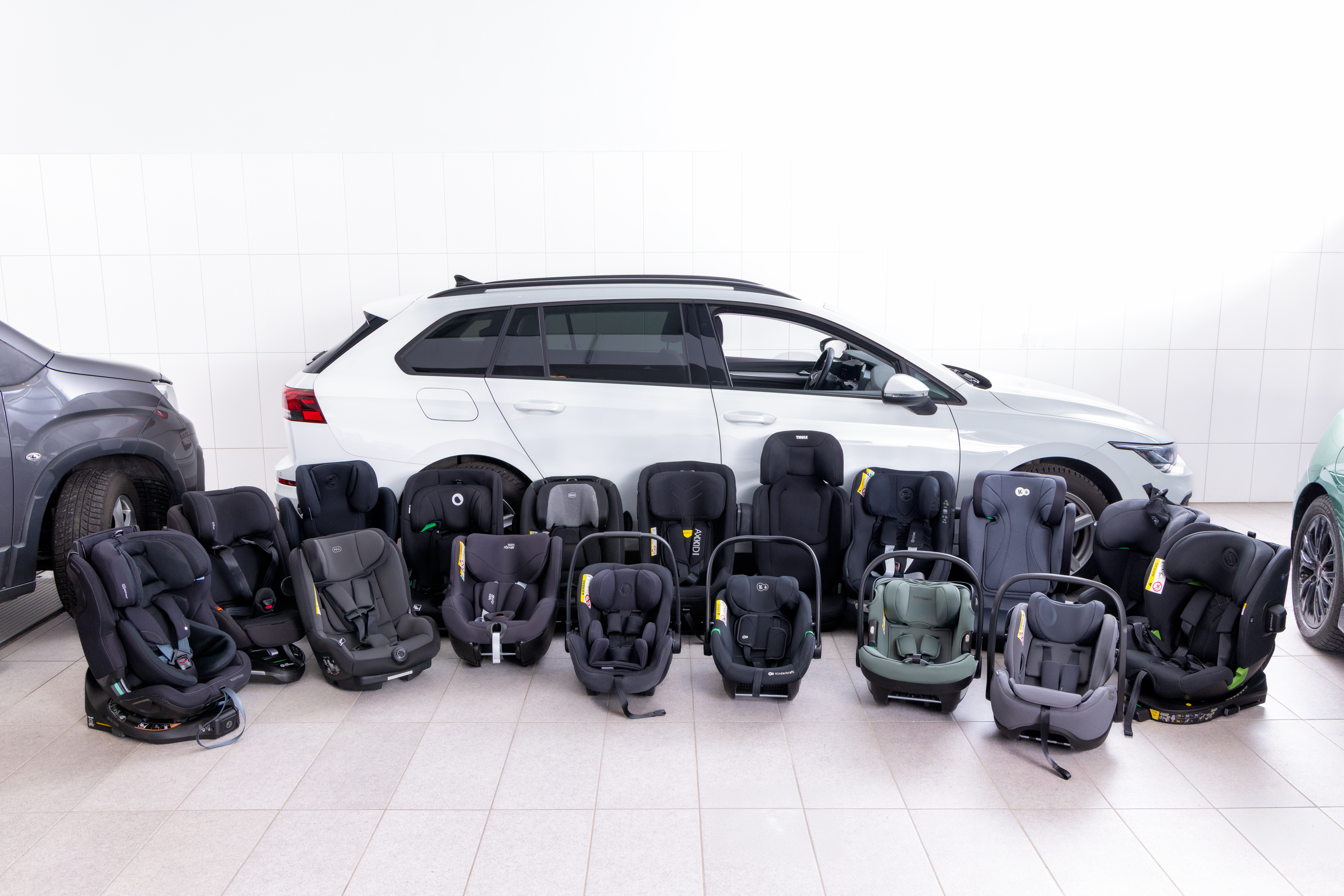 16 Kindersitze stehen nebeneinander am Boden vor einem weißen Auto 