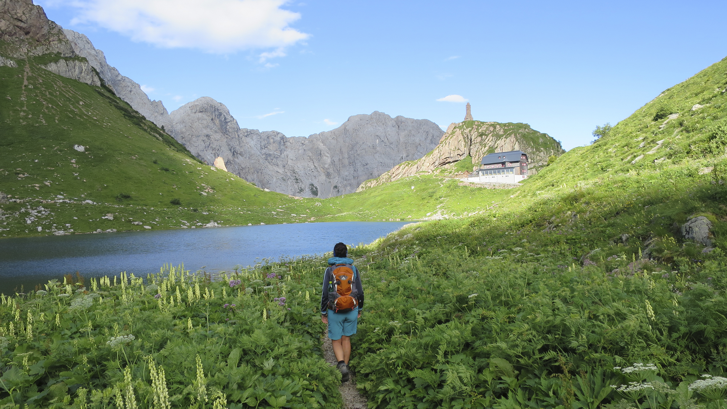 Eine Wanderin geht zu einer Hütte in den Bergen, vorbei an einem Bergsee.