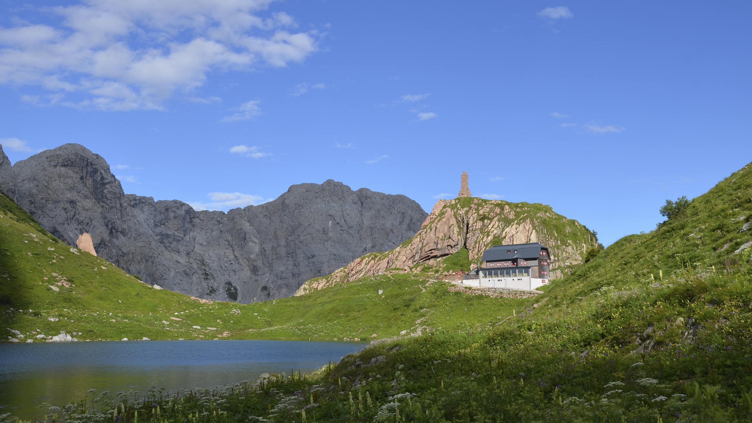 Eine Hütte in den Bergen, im Vordergrund ein kleiner Bergsee.