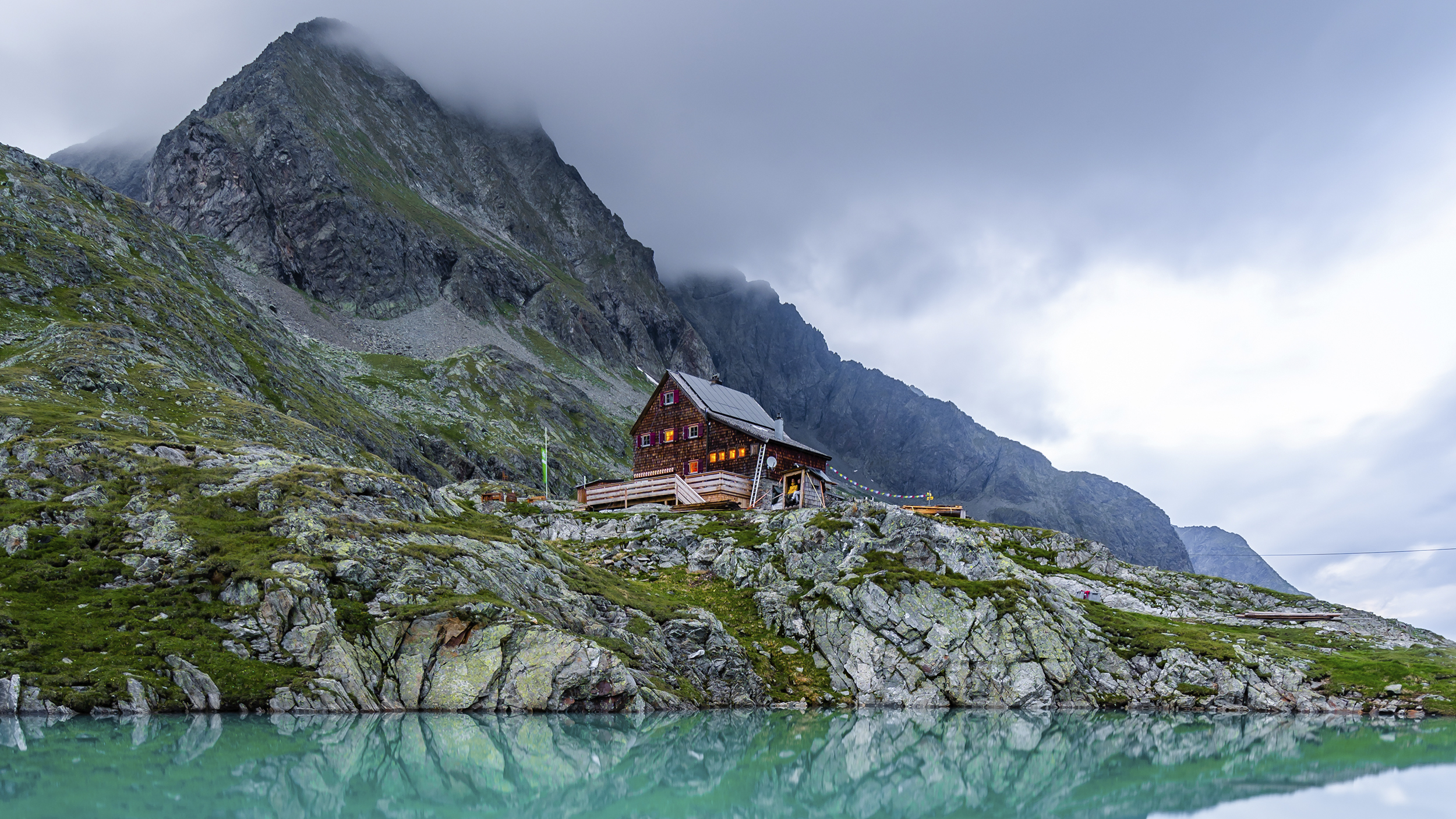 Eine Hütten in den Bergen, im Vordergrund ein kleiner Bergsee.